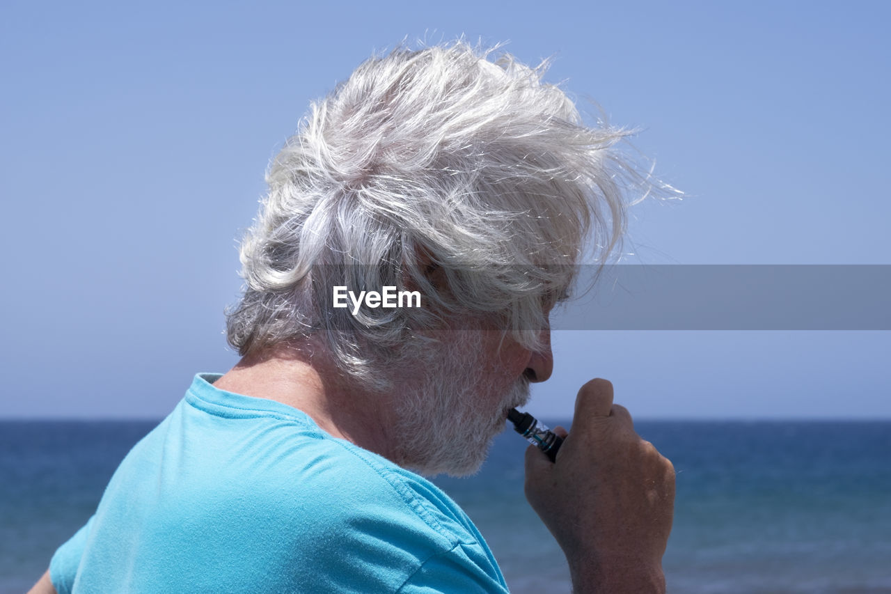 Side view of senior man smoking electronic cigarette against sky