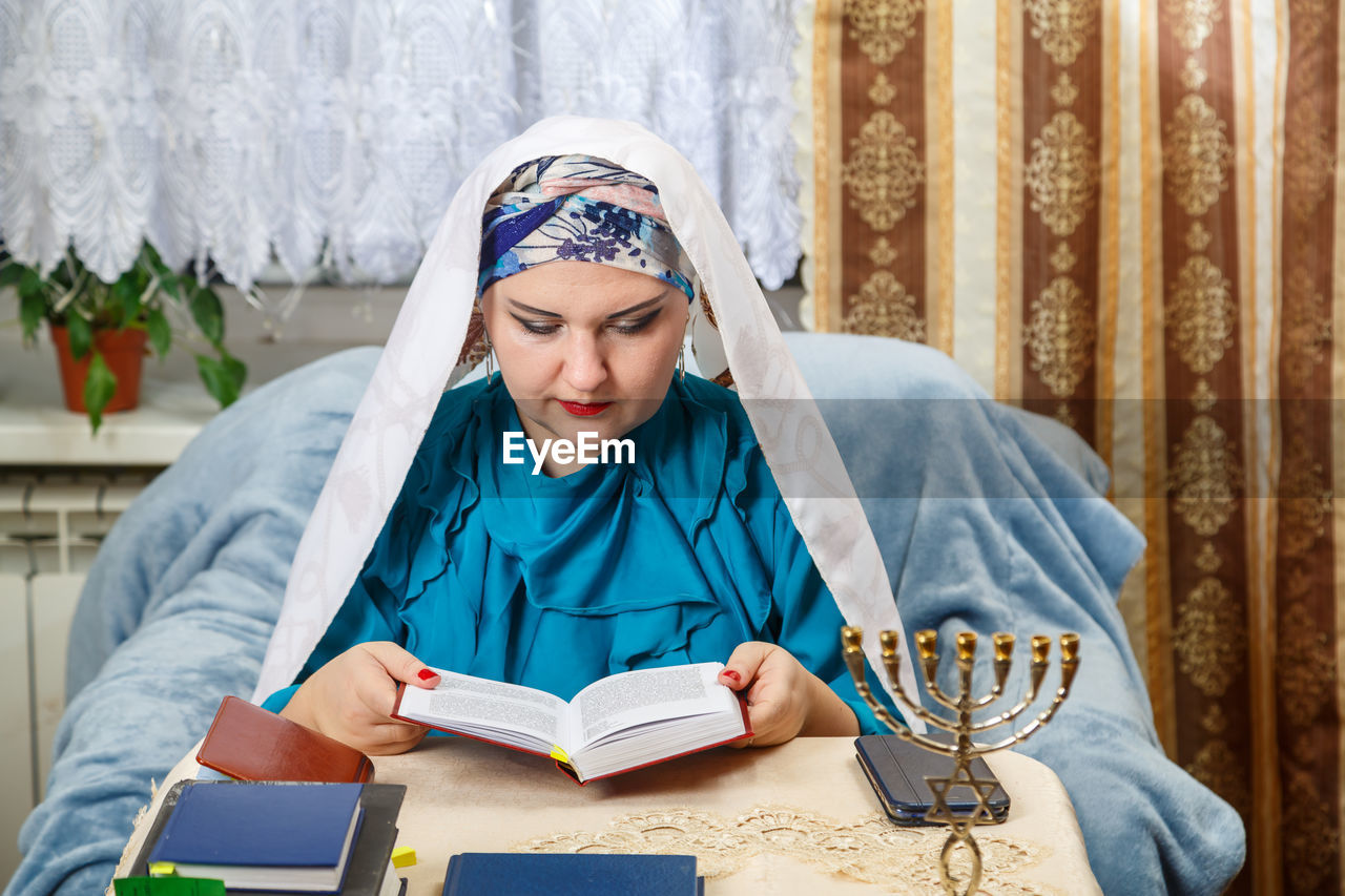 one person, adult, publication, book, person, front view, sitting, women, indoors, headscarf, reading, lifestyles, clothing, looking, young adult, portrait, religion, activity, female, blue, holding, traditional clothing, architecture, furniture, education, belief, looking down, emotion