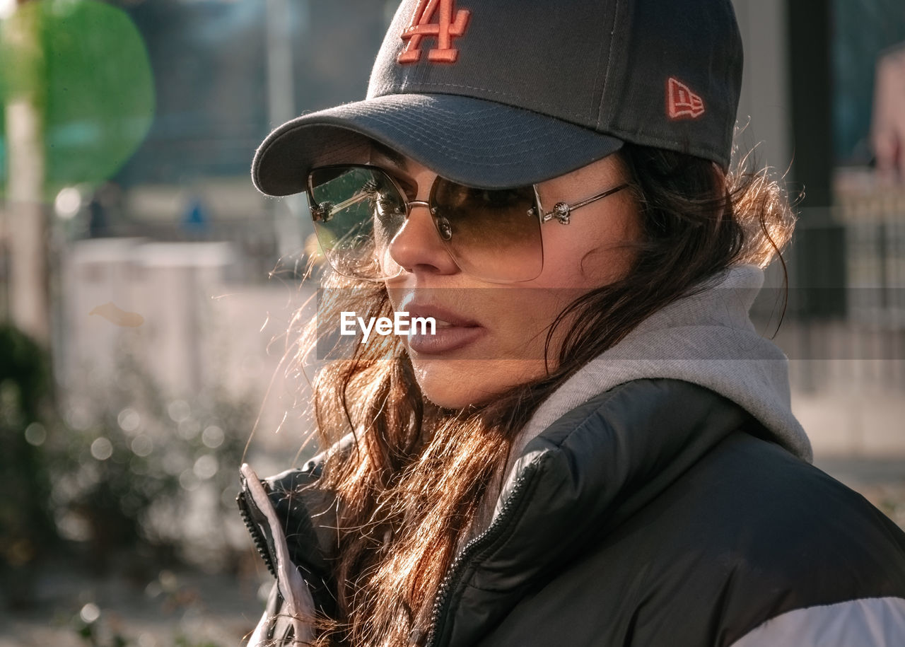 adult, one person, headshot, portrait, clothing, glasses, sunglasses, hat, fashion, women, spring, young adult, day, focus on foreground, hairstyle, cap, outdoors, lifestyles, nature, long hair, leisure activity, city, person, side view, jacket, looking, female, architecture, baseball cap, cool attitude, looking away