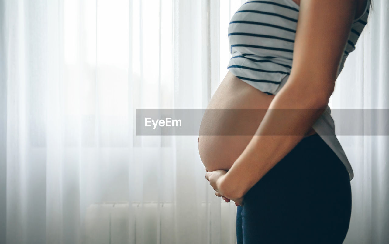 Midsection of pregnant woman touching her belly against curtain at home