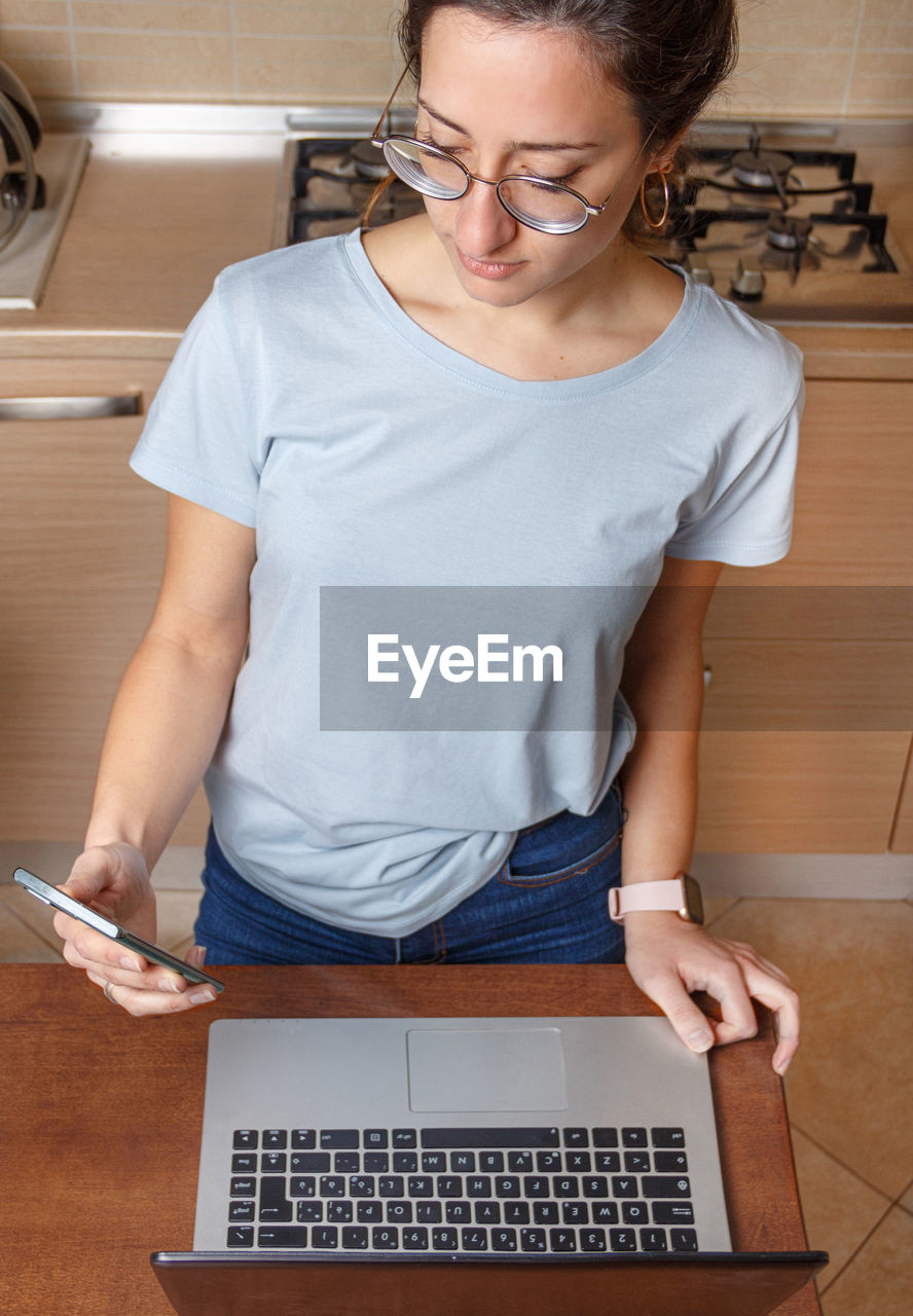 Young women in glasses and blue t-shirt using cell phone and laptop in kitchen