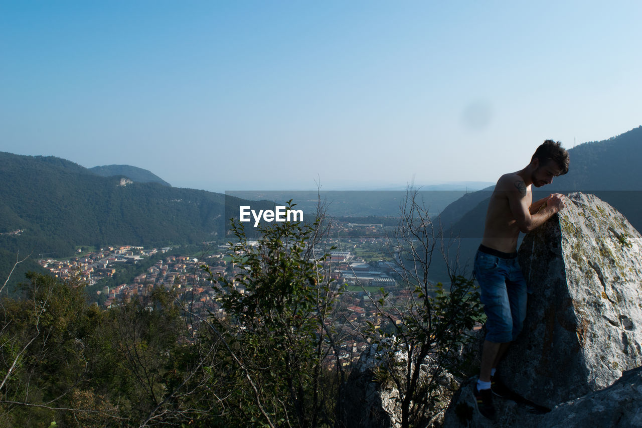 Shirtless young man climbing on rock formation against clear blue sky