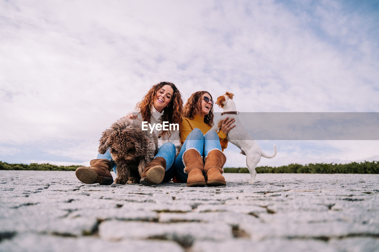 Smiling women playing with dog sitting against sky