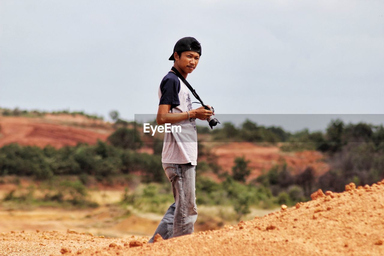 Young man with camera standing on hill against sky