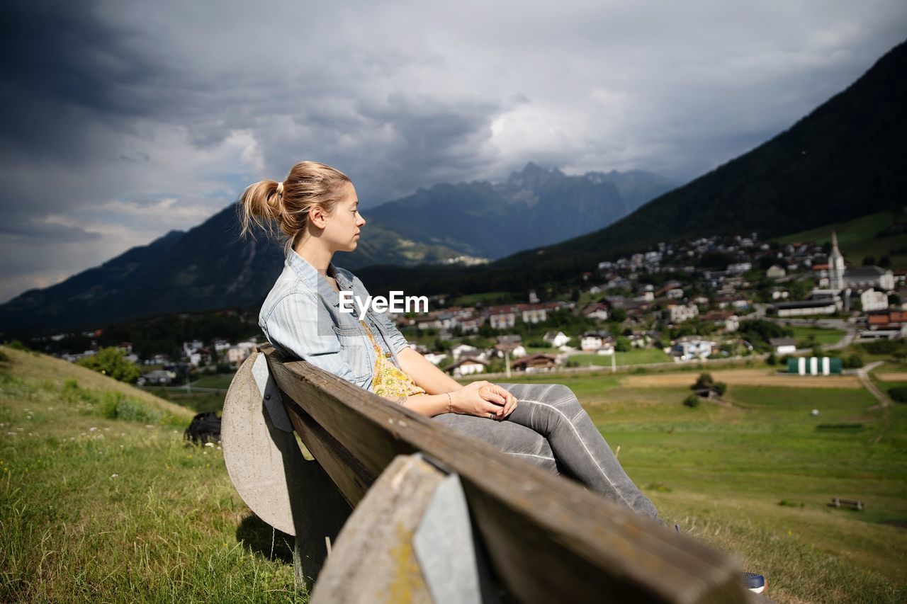 Woman looking away while sitting on bench against cloudy sky