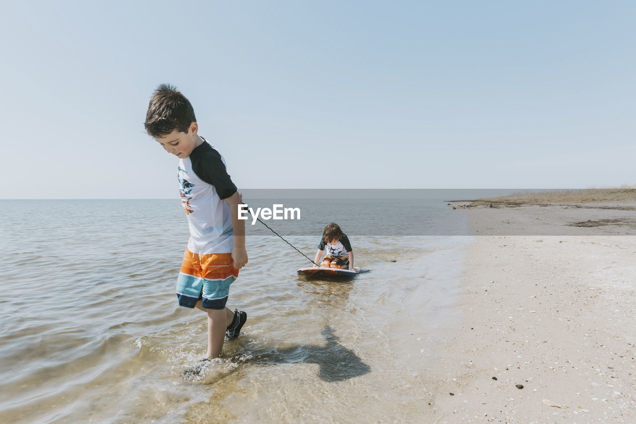 Boy pulling brother sitting on surfboard in sea against clear sky during sunny day