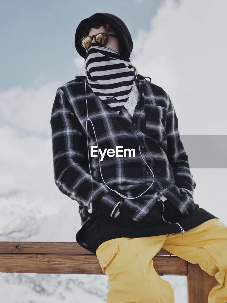 Low angle view of man sitting on railing outside snow against sky