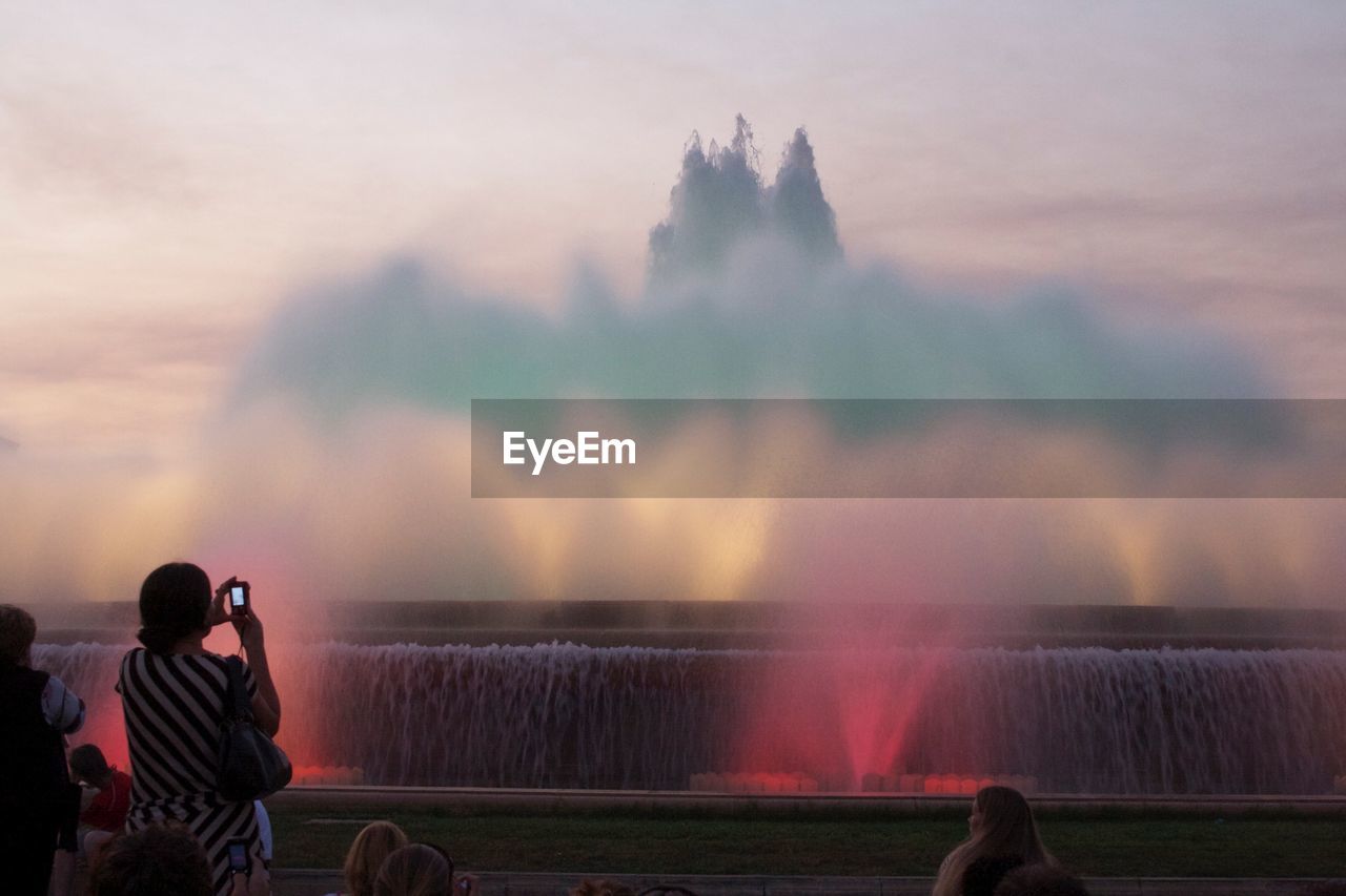 Rear view of woman photographing colorful fountain during sunset