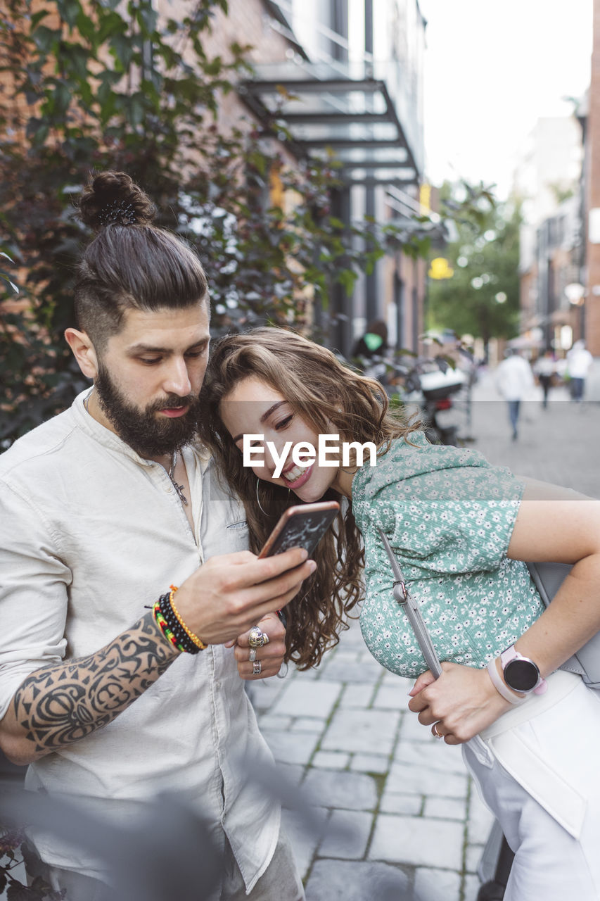 Hipster man showing smart phone to girlfriend while standing on footpath