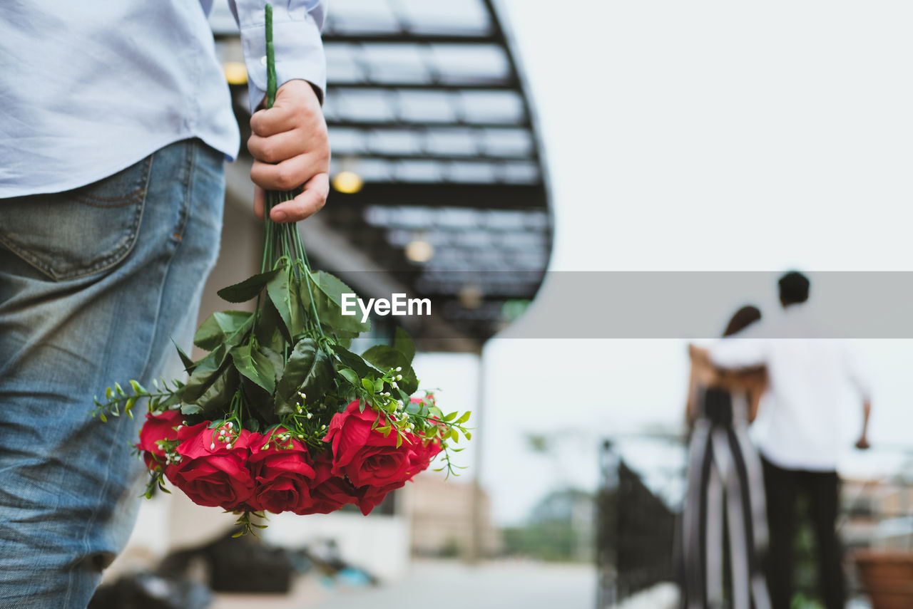 Midsection of man holding rose bouquet while couple standing against clear sky