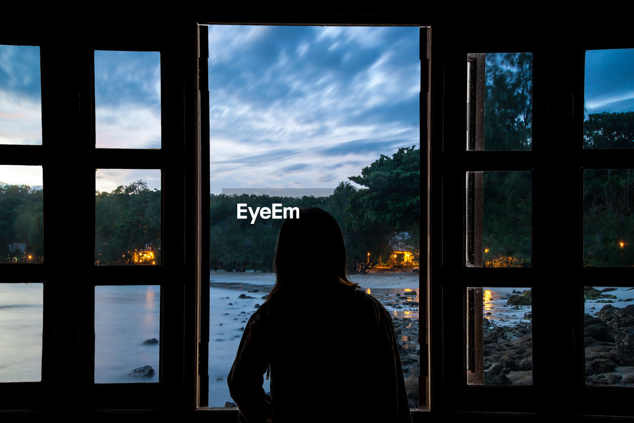 Woman looking at lake through window during sunset