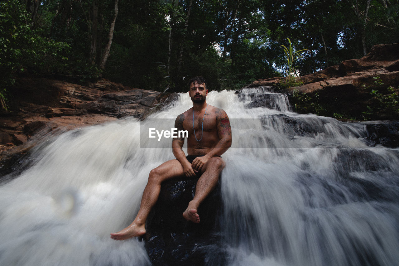Full length portrait of shirtless man sitting on rock amidst waterfall