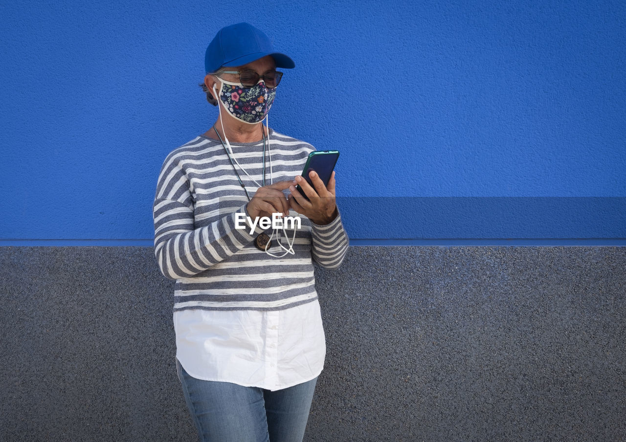 Senior woman wearing mask using smart phone standing by wall