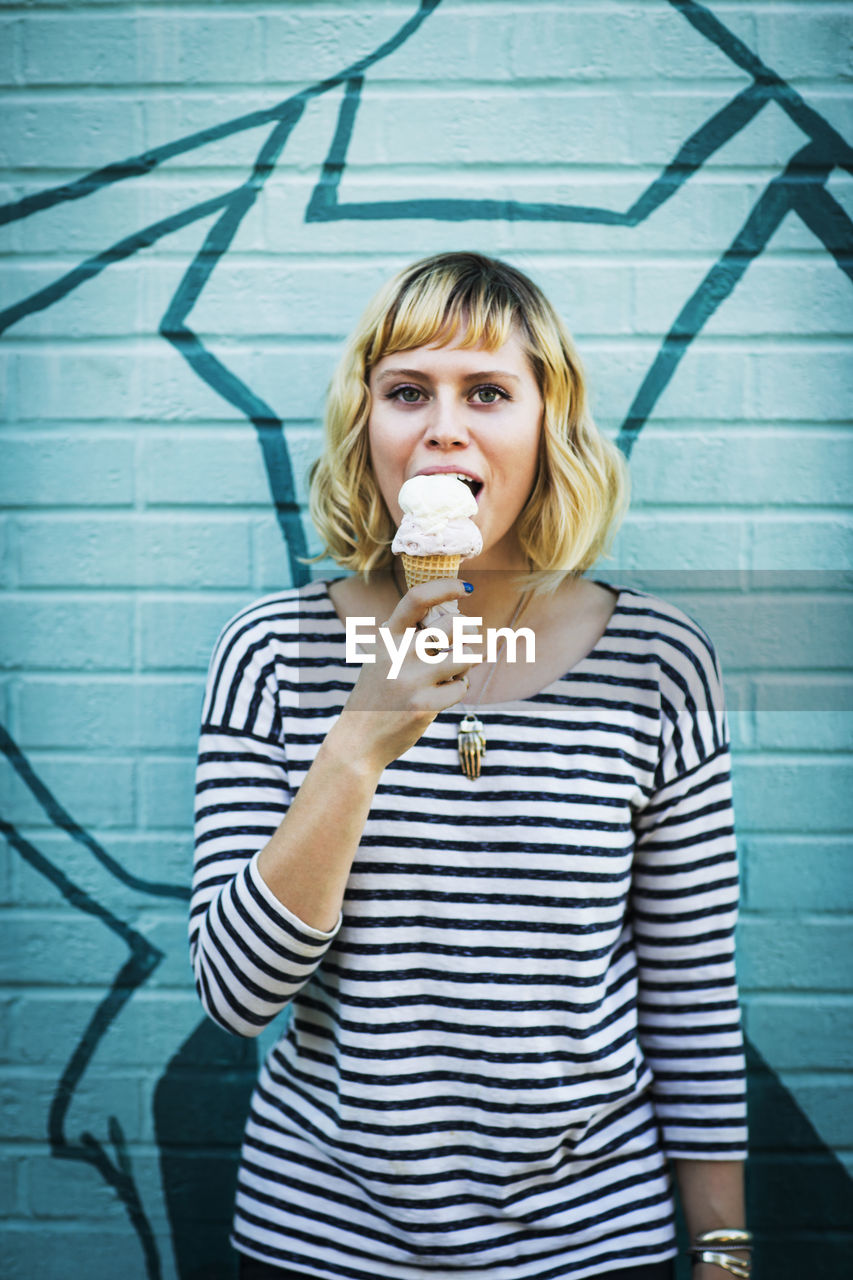 Portrait of woman eating ice cream while standing against wall