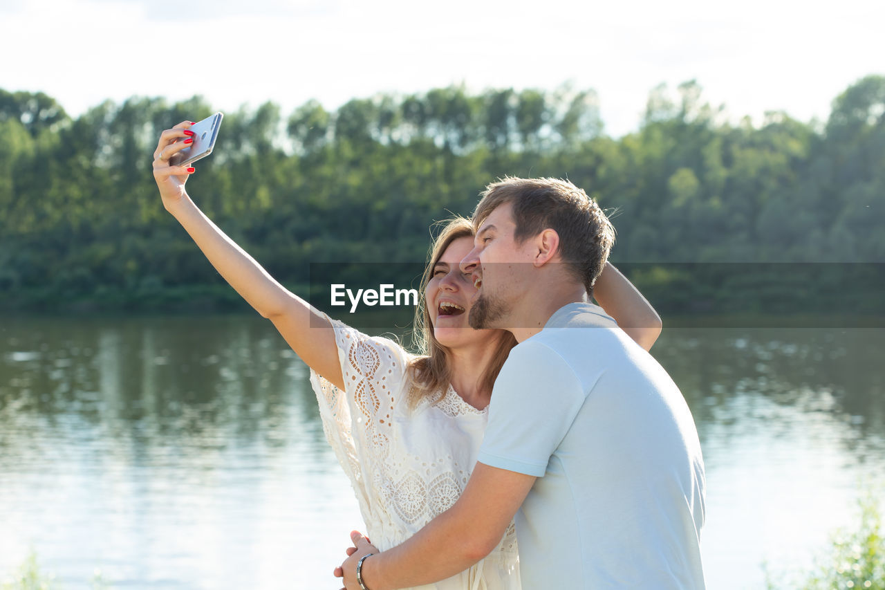 Young couple doing selfie while standing against lake