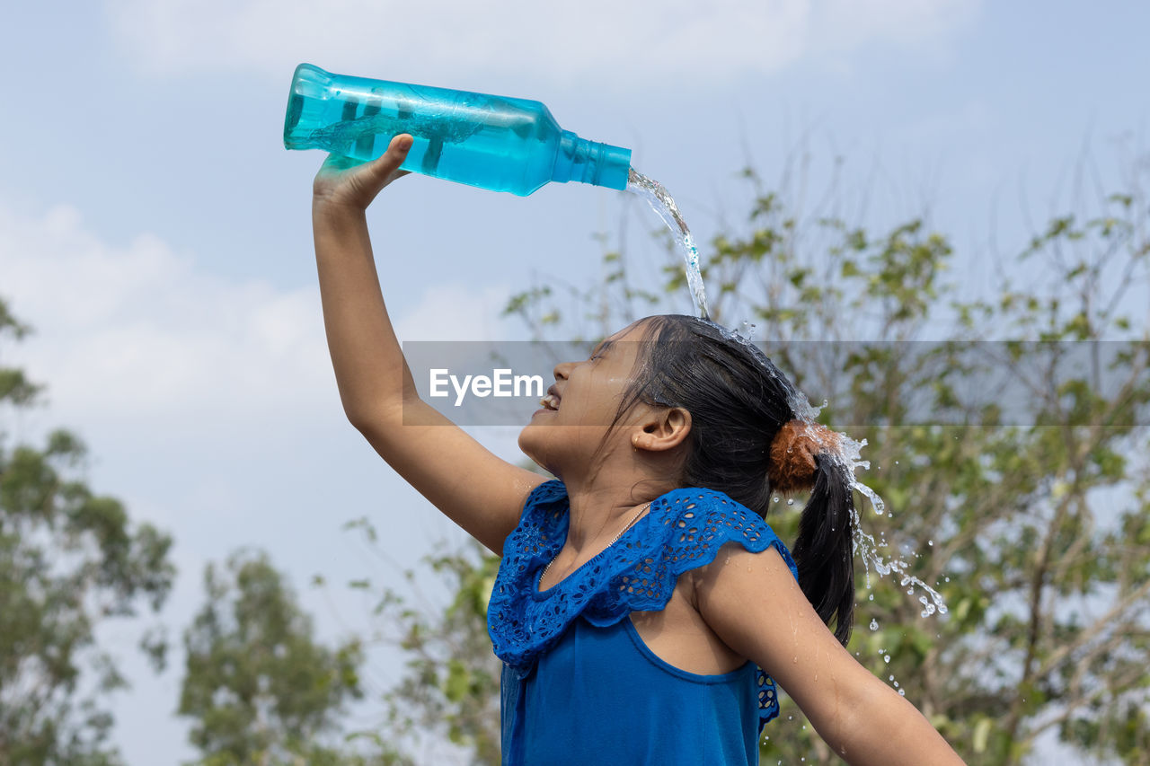 An indian girl pouring water on head from a blue plastic bottle under sky to cool down in summer