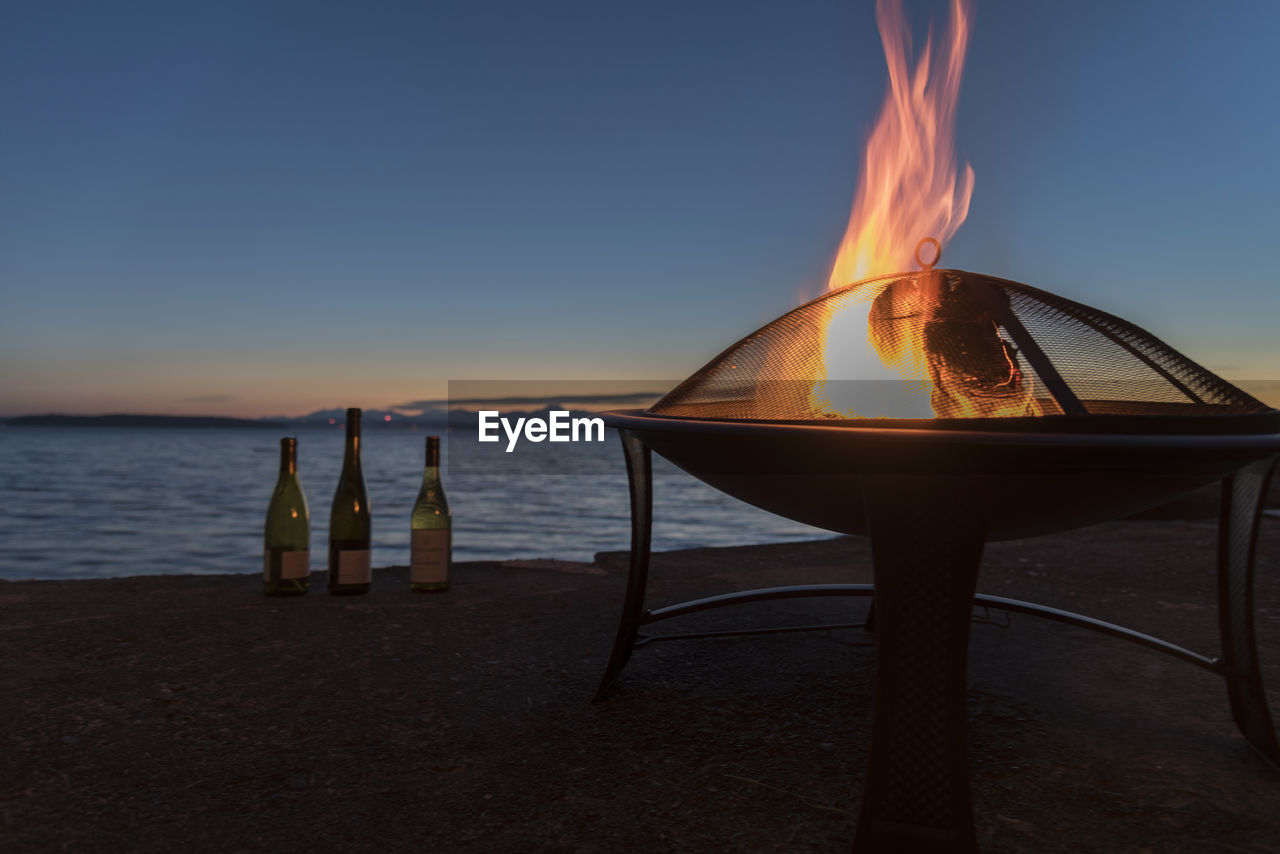 Wine bottles by burning fire pit at beach against sky during sunset