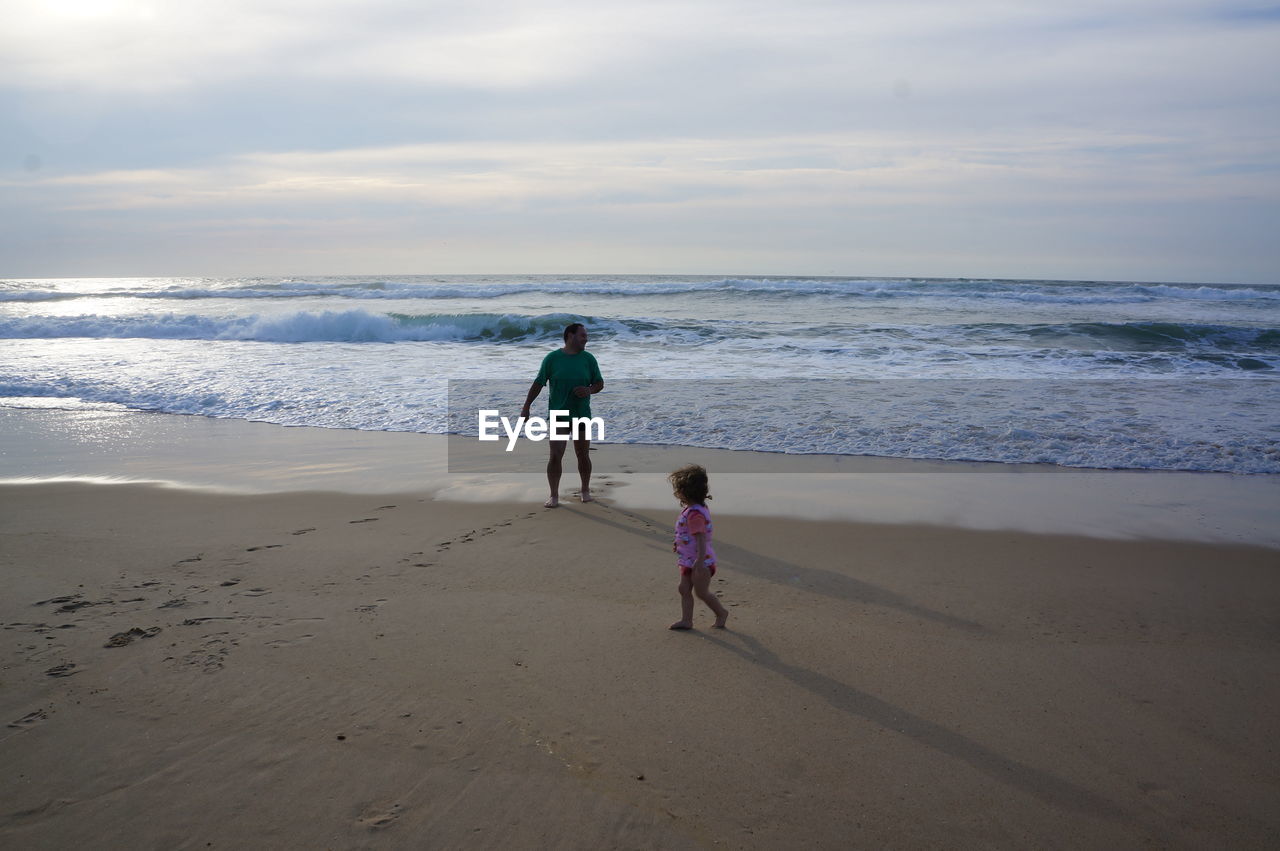 Full length of father and daughter walking at beach against sky