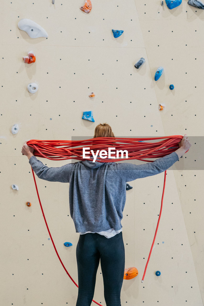 Back view of sportive female with ropes for climbing behind back standing near wall during training in gym