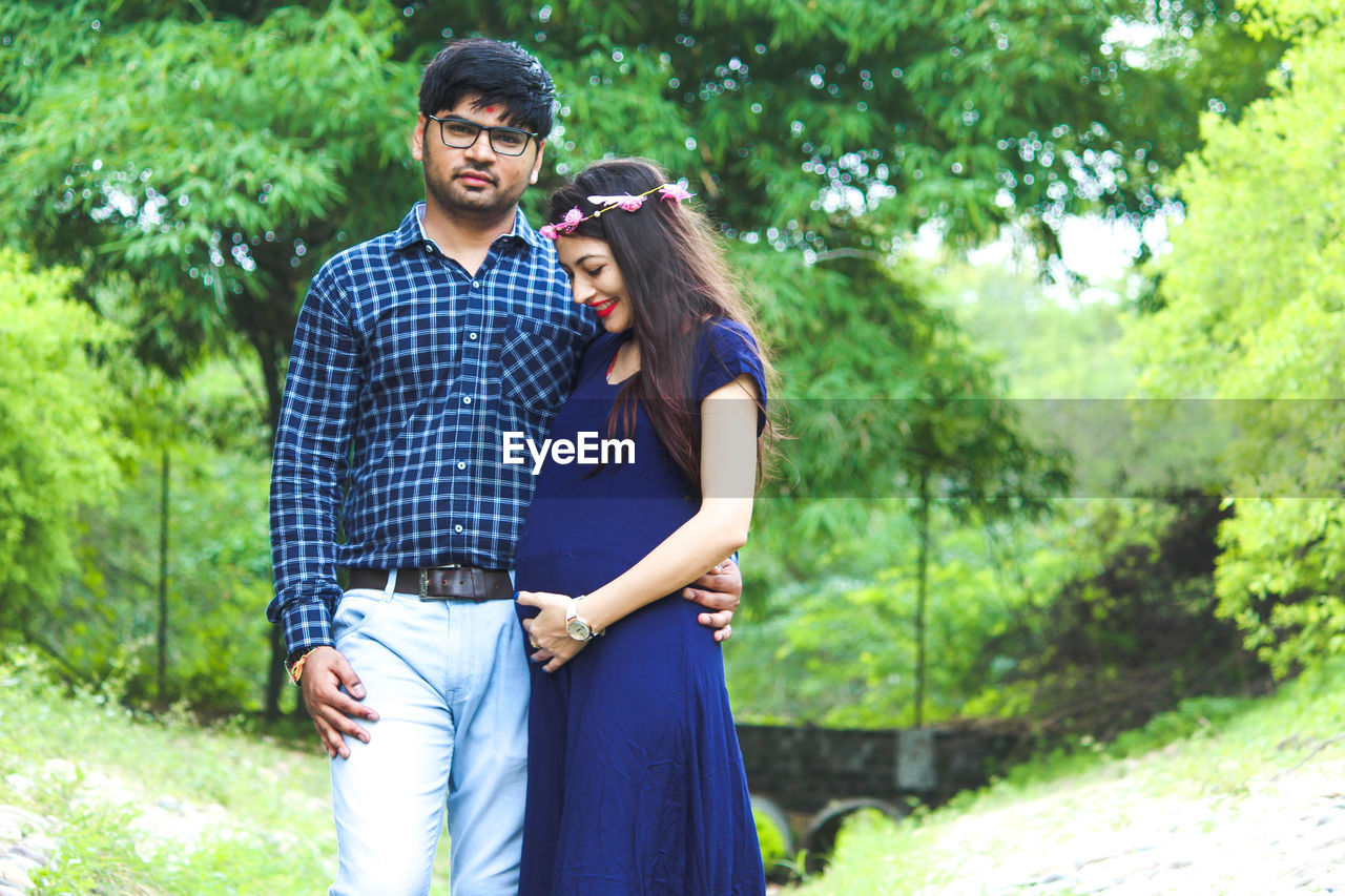 Portrait of young man standing with pregnant woman against trees