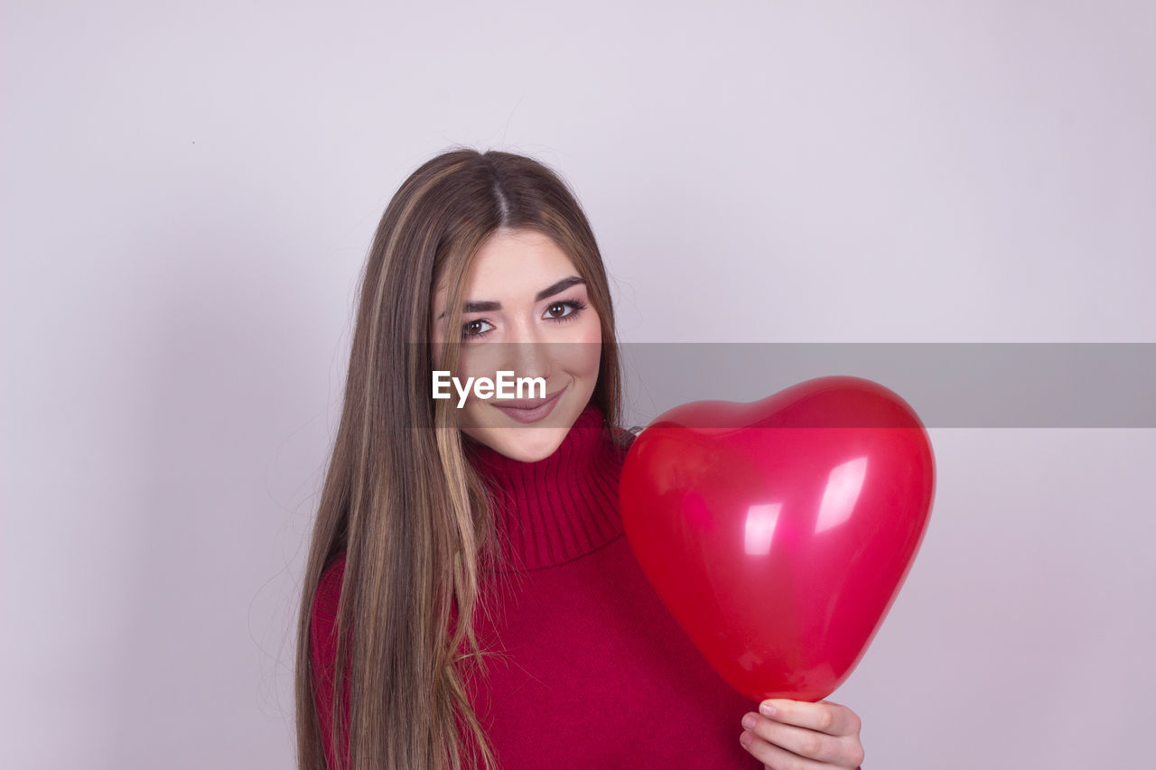 Portrait of smiling young woman holding heart shape balloon against colored background