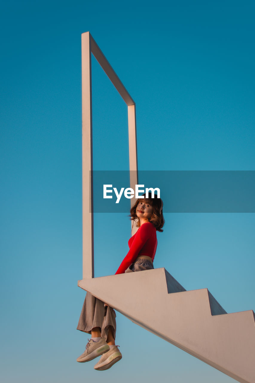 LOW ANGLE VIEW OF BOY STANDING BY STAIRCASE AGAINST CLEAR BLUE SKY