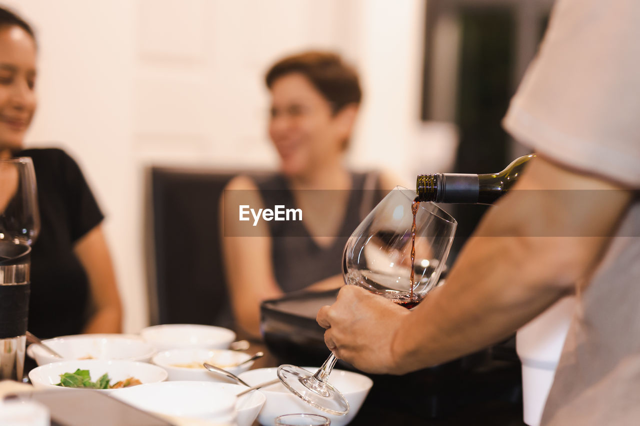 Selective focus of man pouring red wine with woman friend smiling in background.