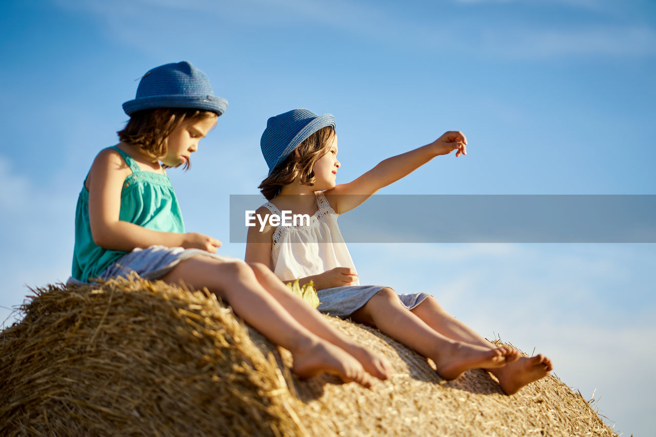 Low angle view of friends wearing hats sitting on hay bale against sky