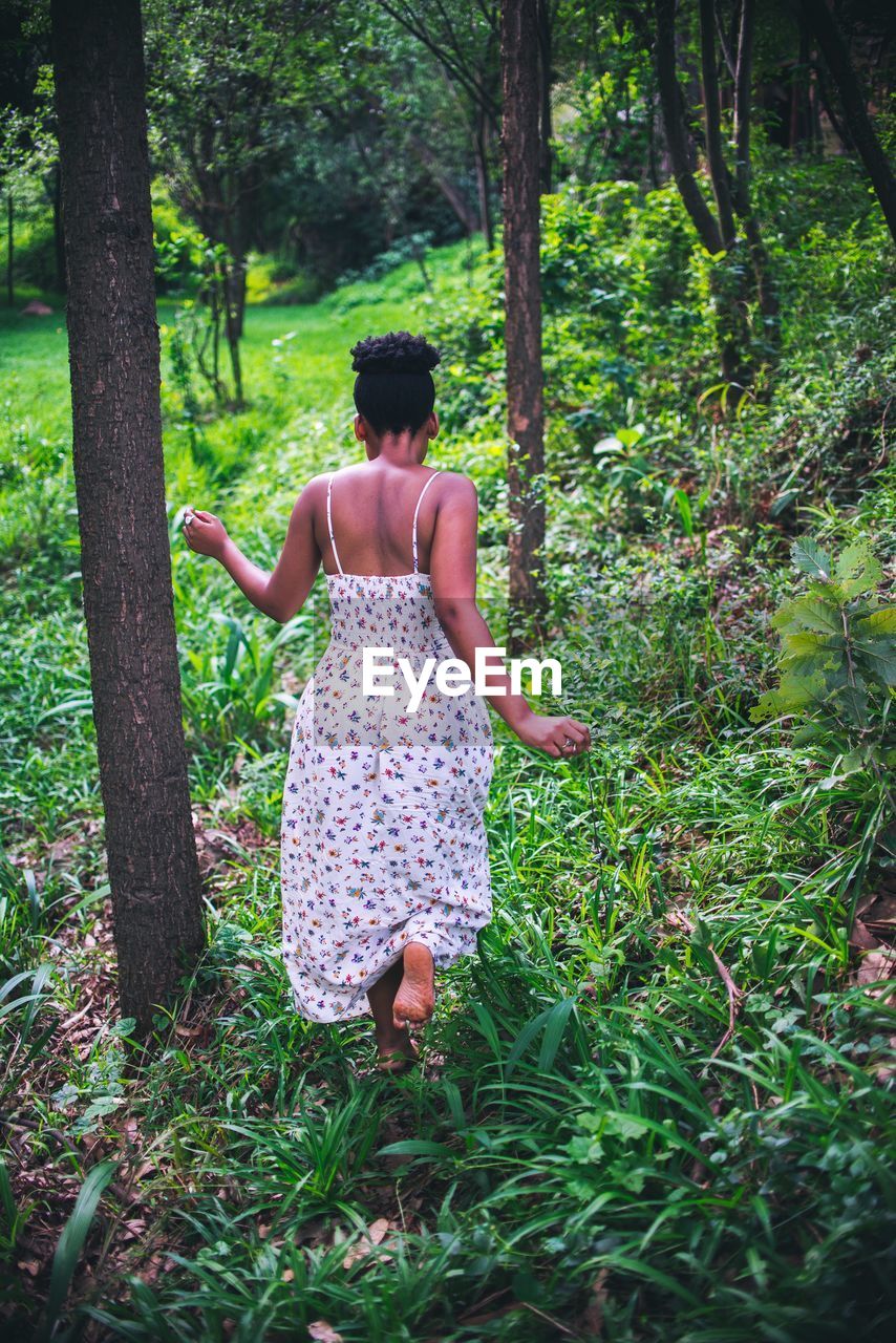 REAR VIEW OF WOMAN WALKING ON DIRT ROAD AMIDST TREES