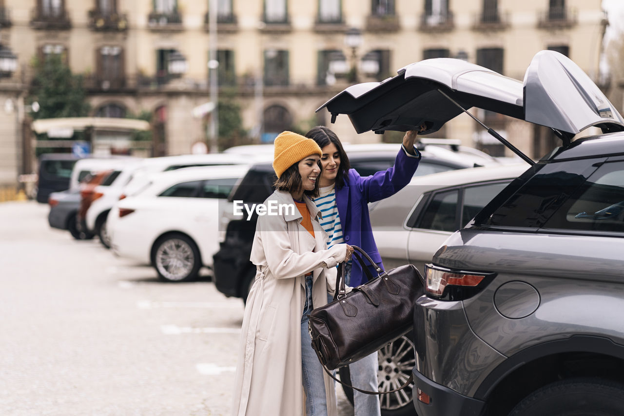 Positive female friends in stylish wear putting bag into truck of modern automobile parked on street of city with building