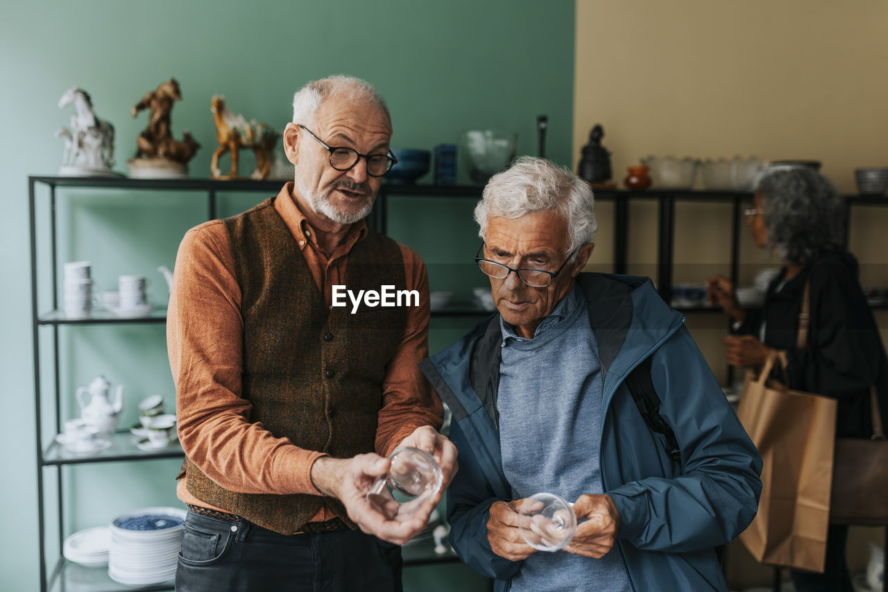 Senior male entrepreneur showing drinking glass to customer in antique shop