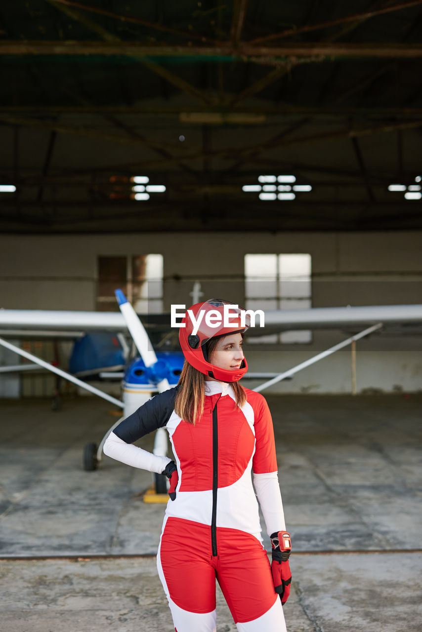 Young female skydiver in a plane hangar with a plane behind her