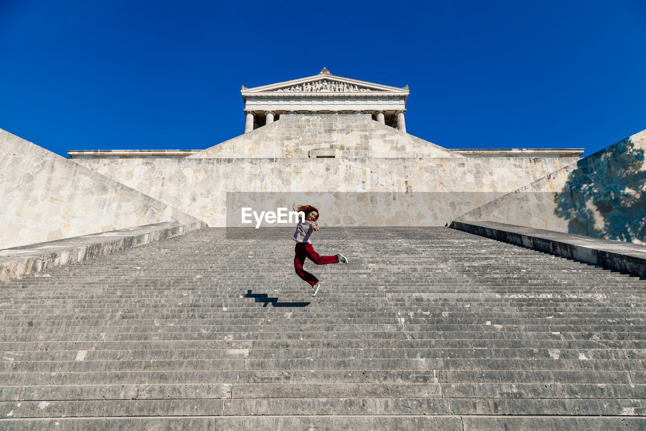 Low angle view of happy woman jumping on staircase against clear blue sky