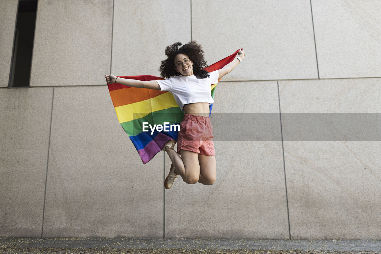 Cheerful woman jumping while holding rainbow flag in front of wall
