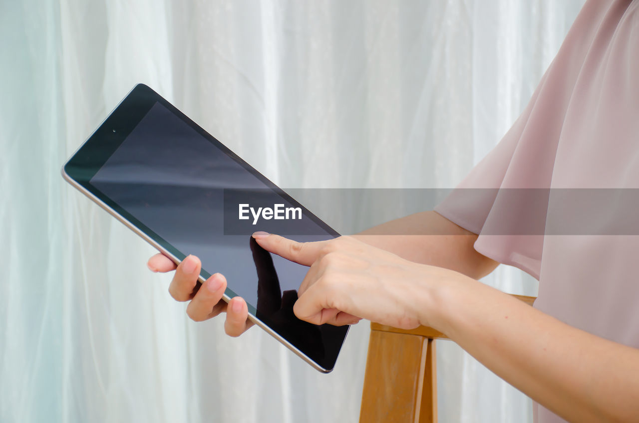 Midsection of woman using digital tablet against curtain