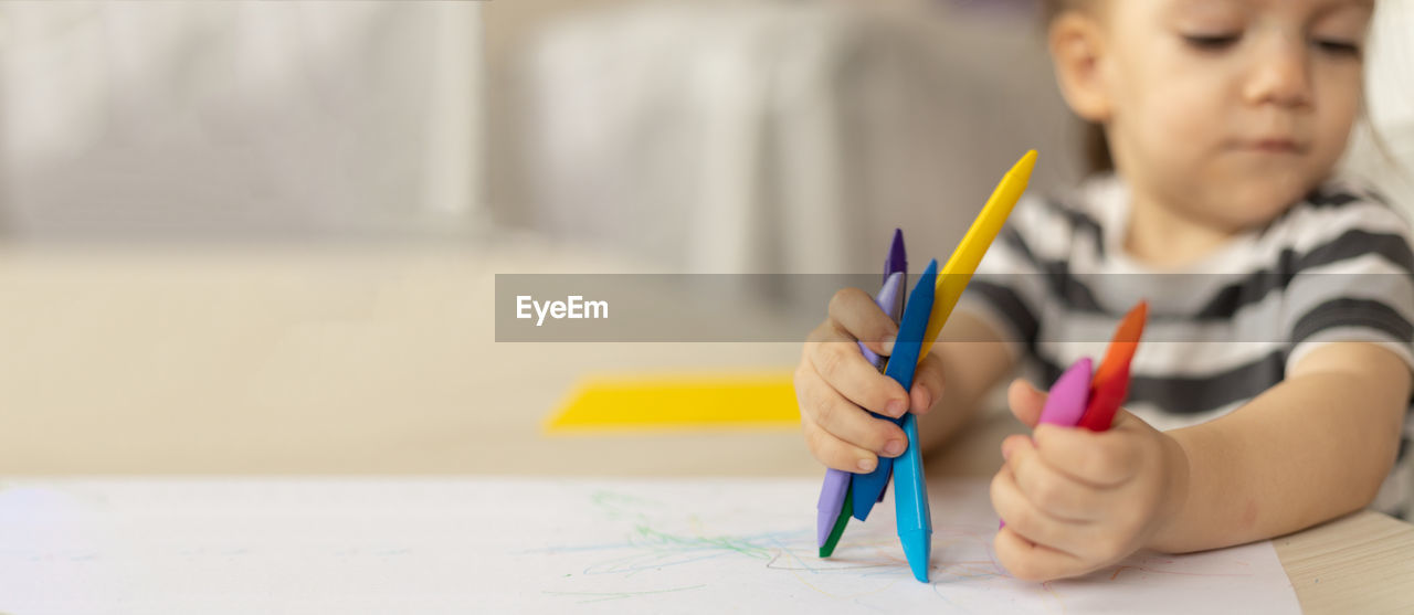 Banner cute caucasian baby girl holding wax crayons in hands sitting at the table at home drawing