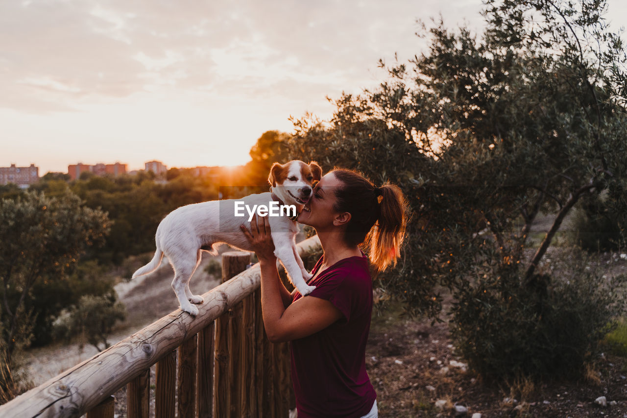 Woman holding dog against sky during sunset