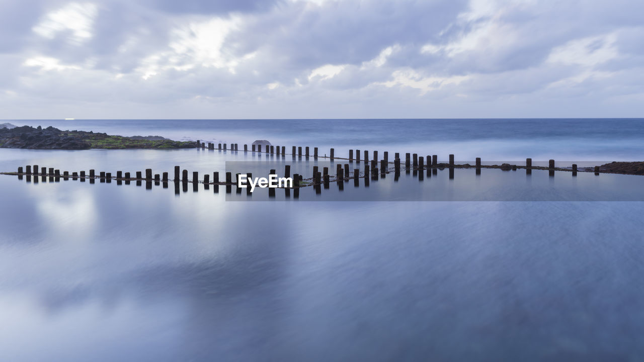 Wooden posts in sea against cloudy sky