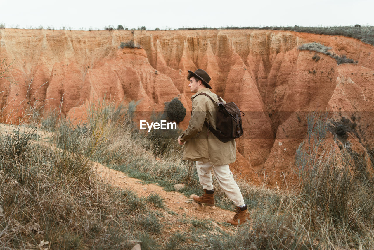 Side view of young man in casual clothes with backpack and hat walking on path near arid canyon during trip in countryside