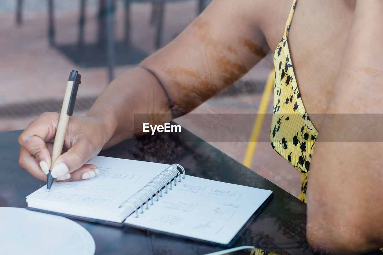 Crop view of black self employed woman sitting at table in cafe and working remotely while writing in notepad