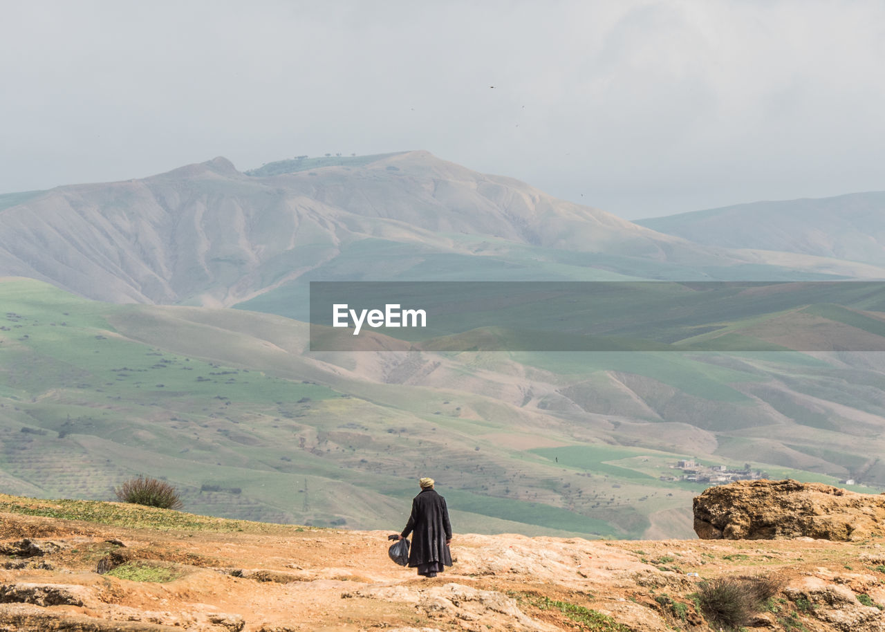 REAR VIEW OF MAN ON LANDSCAPE AGAINST MOUNTAINS