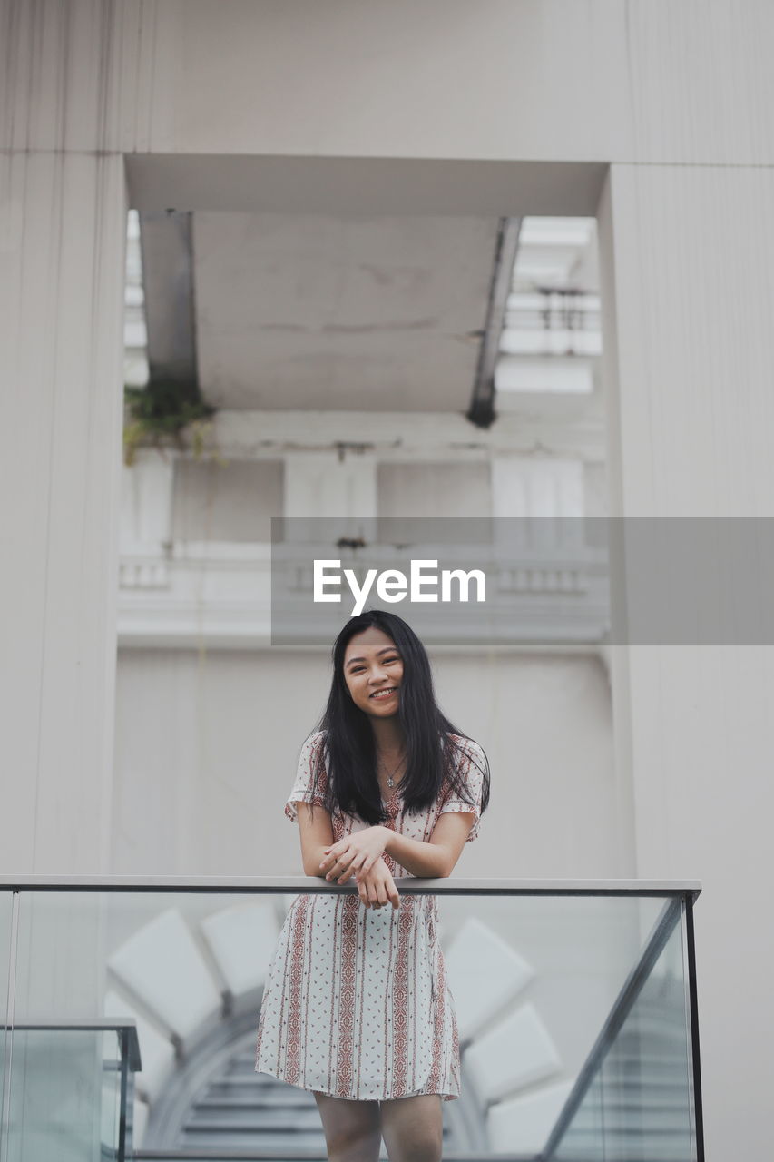 Portrait of young woman standing by railing against building 