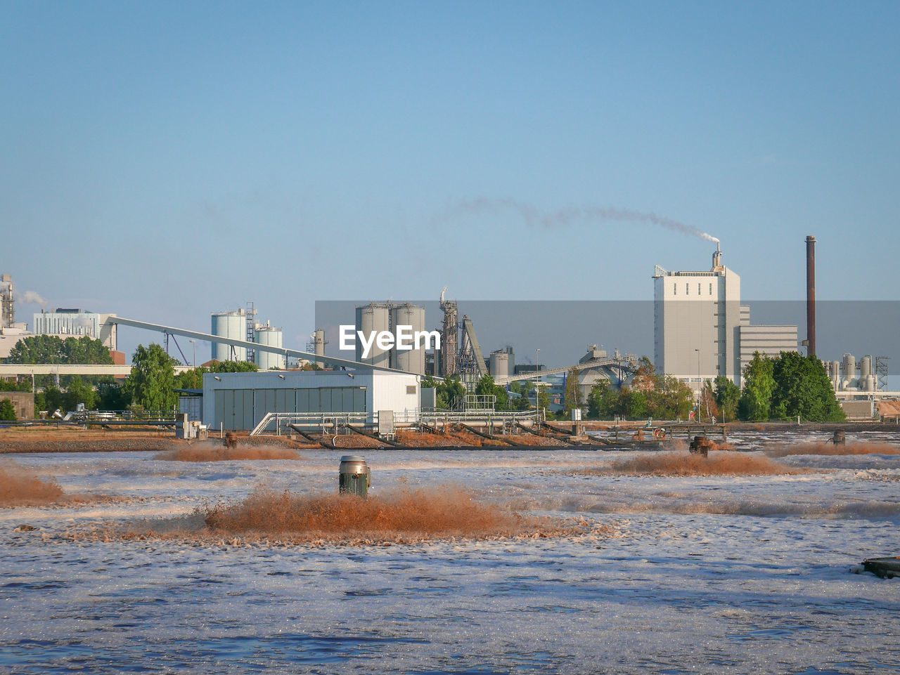 Smoke emitting from factory by river against sky