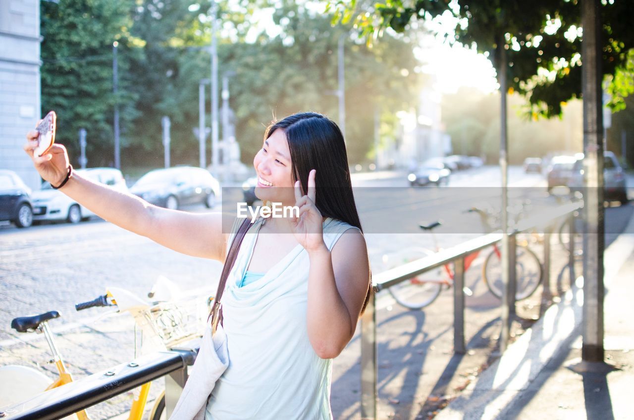 Woman gesturing while taking selfie through mobile phone on sidewalk