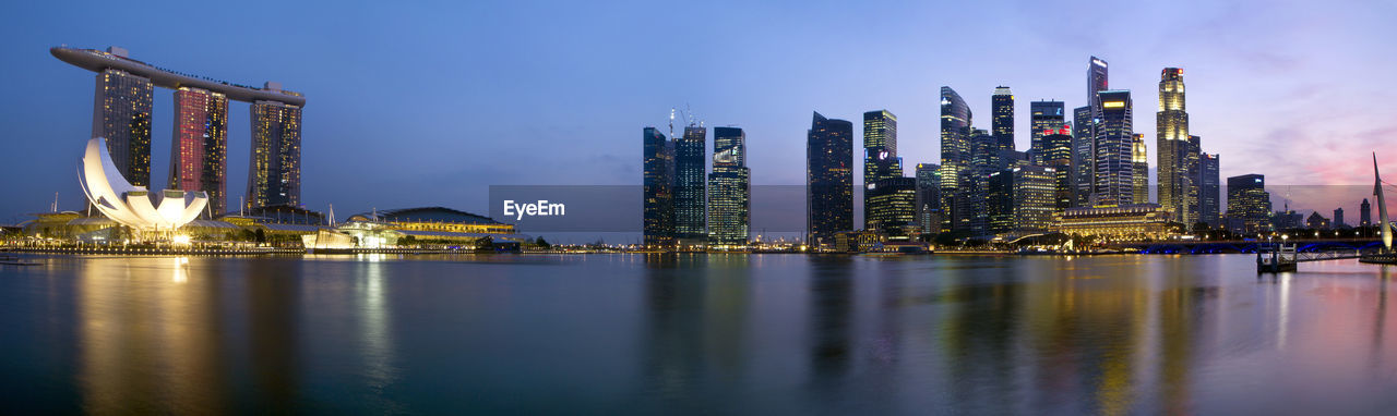 Illuminated modern buildings by river against sky