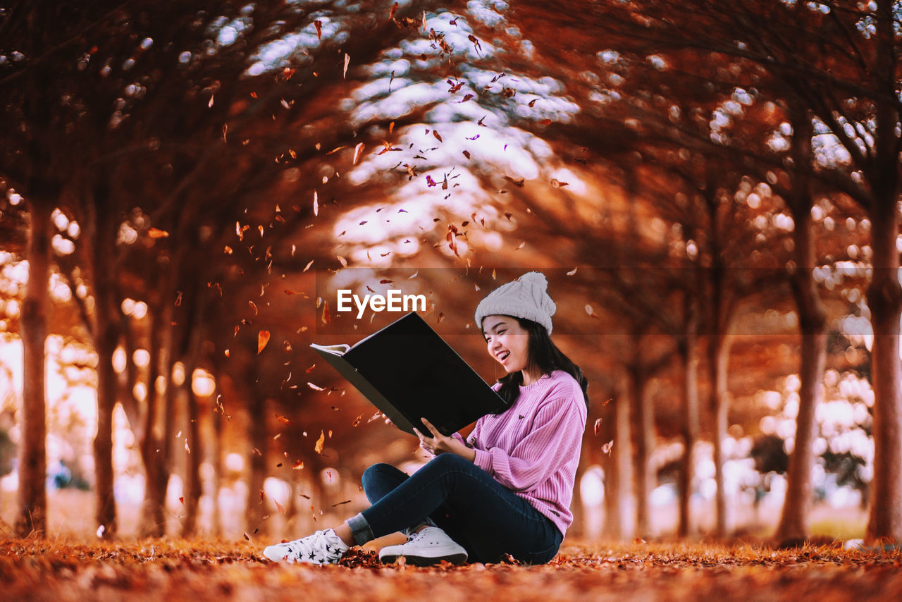 Cheerful young woman reading book while sitting in park during autumn