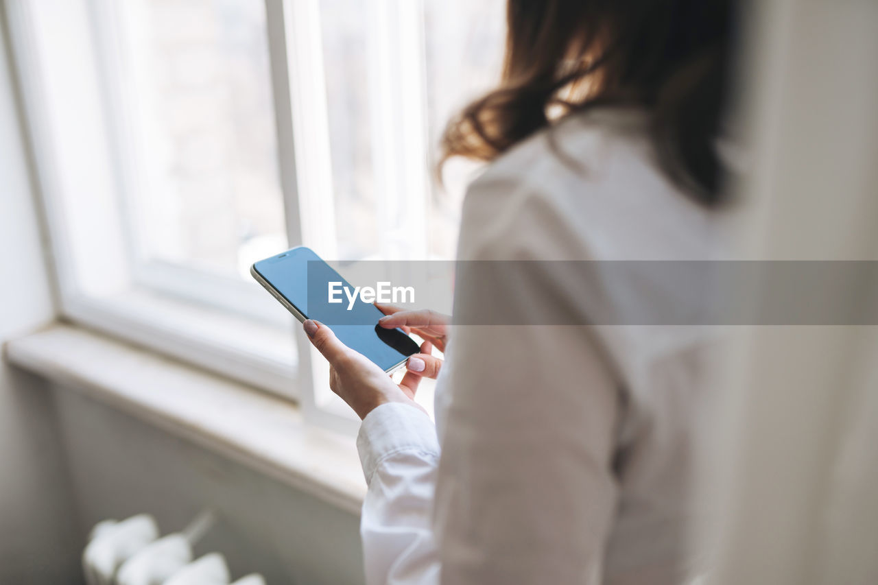 Crop photo of young brunette woman in white shirt using mobile phone in hands near window