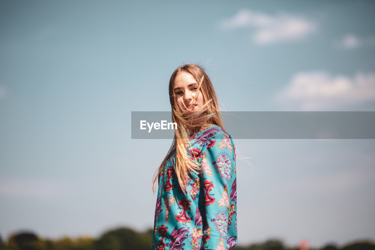 Portrait of female model with tousled hair standing against sky