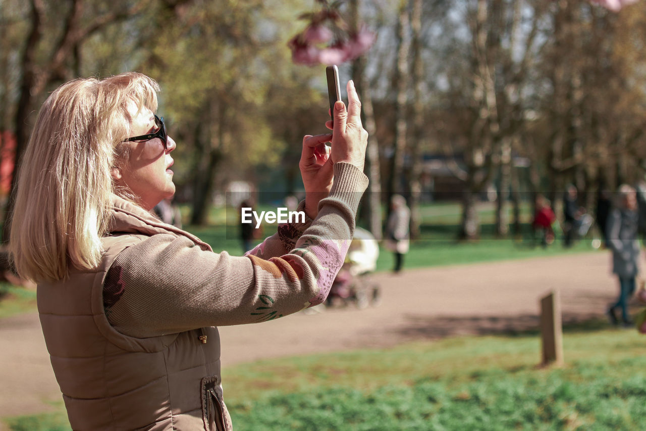 SIDE VIEW OF WOMAN HOLDING SMART PHONE WHILE STANDING ON PLANT