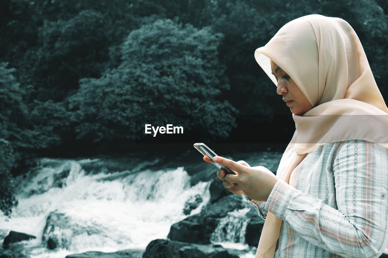 Side view of woman using mobile phone by waterfall in forest